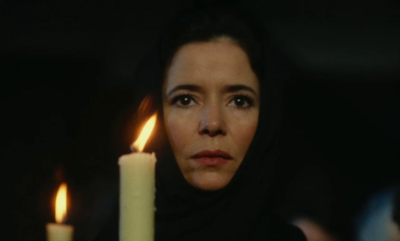 Litan (1982) movie still: closeup — A dark, minimally lit space with a woman standing against a black background. She stares forward while holding a…