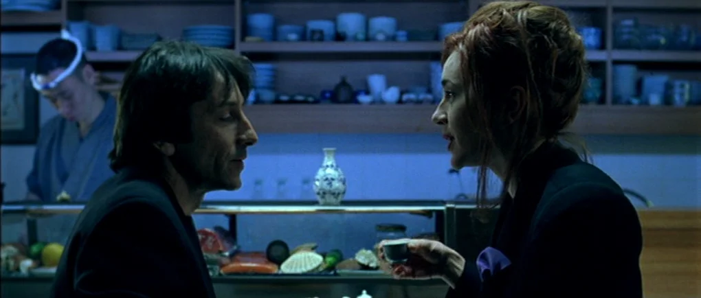 Mortal Transfer (2001) movie still: medium — A dimly lit sushi restaurant interior features shelves of ceramics against a dark back wall. A man and…