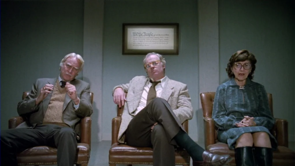 Patty Hearst (1988) movie still: medium — A waiting room with sage green walls featuring a framed document centered behind chairs. Three people sit…