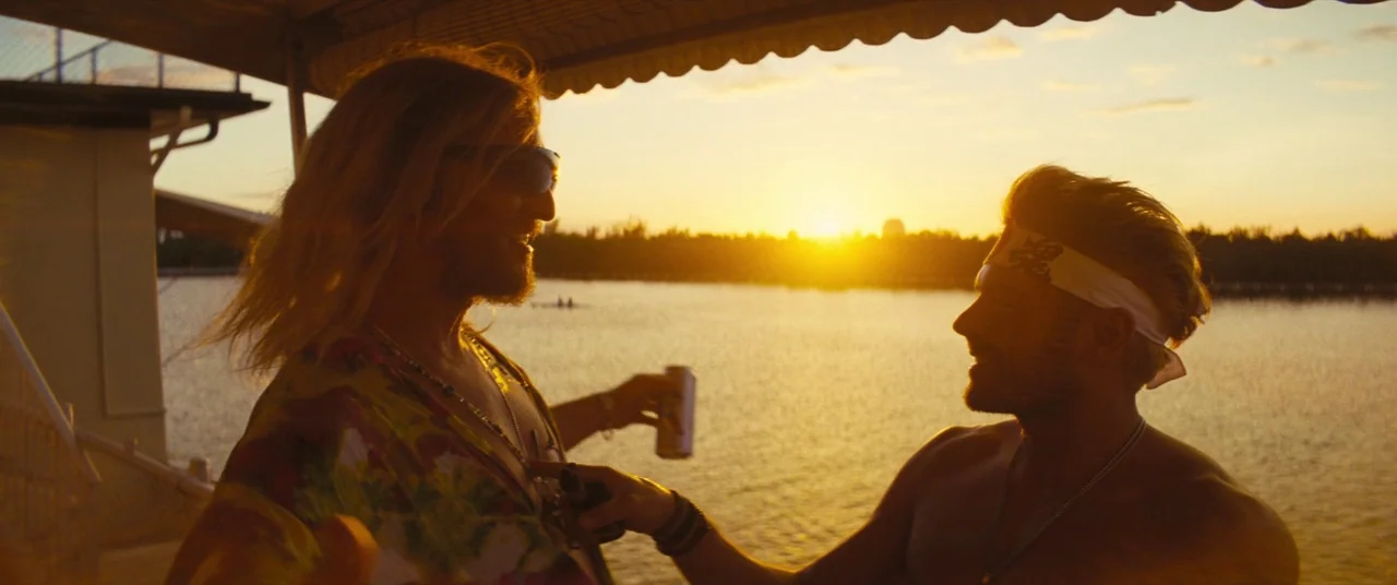 The Beach Bum (2019) movie still: medium — Two men stand on a wooden dock overlooking a lake during a sunset. The men engage in conversation while…