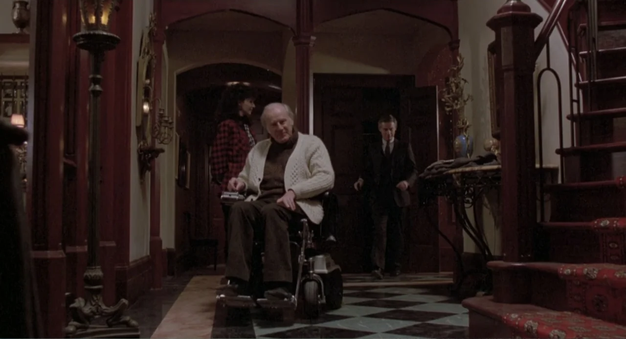 Dead of Winter (2025) movie still: medium — A grand Victorian-style foyer features a checkered marble floor and a wooden staircase. An elderly man…