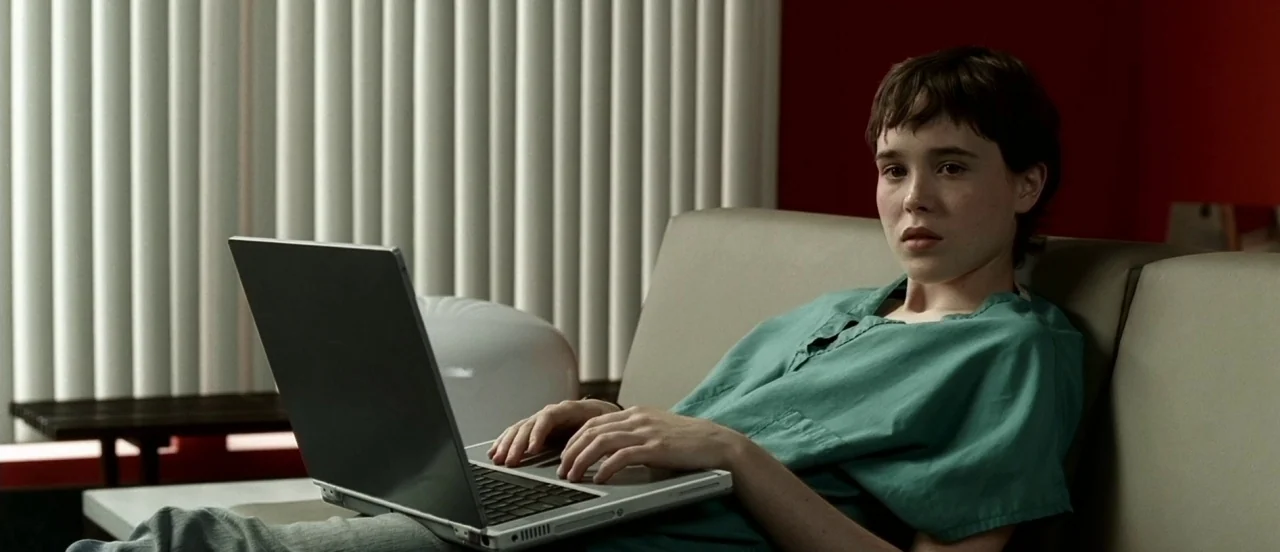 Hard Candy (2005) movie still: medium — A quiet indoor space features vertical blinds over a window and a red wall. A young woman in medical scrubs…
