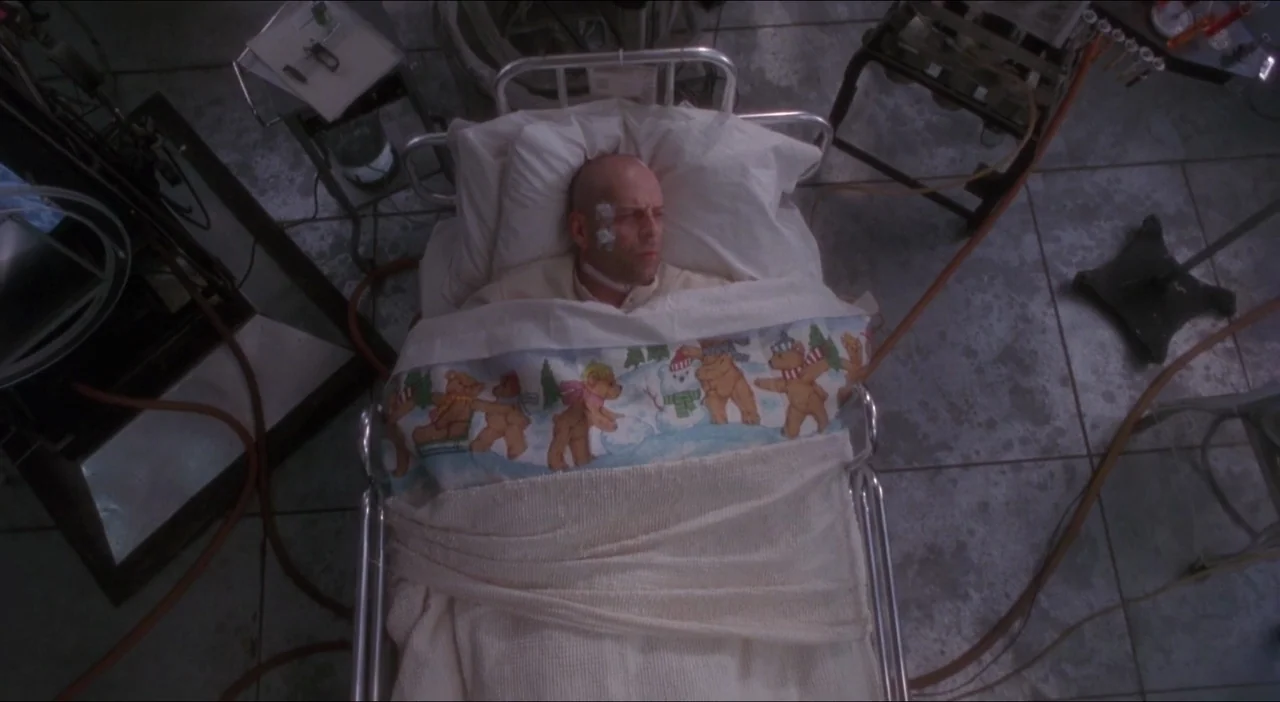 Twelve Monkeys (1995) movie still: wide — A clinical tiled hospital room contains a metal bed and scattered medical apparatus. A man rests in the…