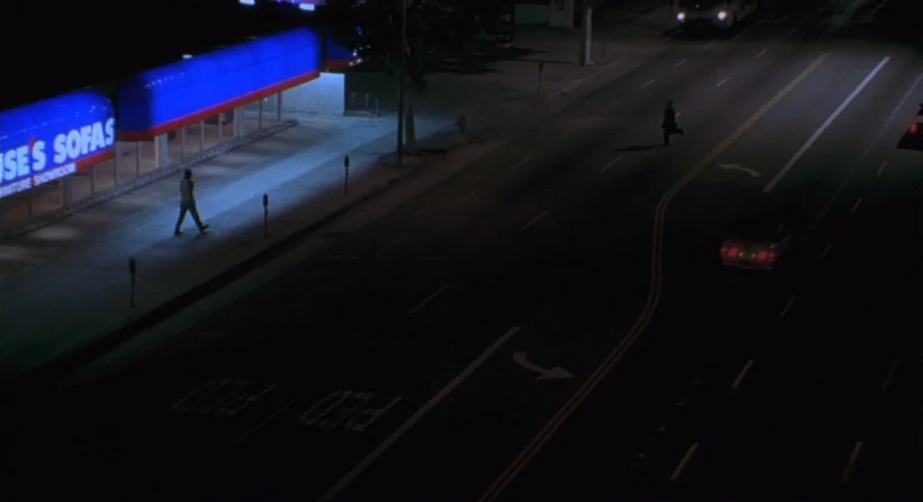 The Crossing Guard (1995) movie still: wide — A night view of a multi-lane asphalt road adjacent to a furniture showroom. Two individuals are moving…