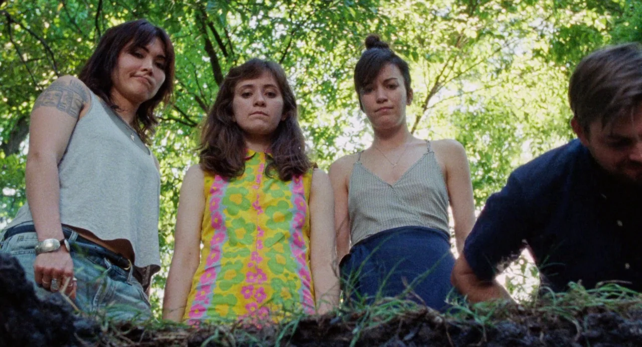Mr. Roosevelt (2017) movie still: medium — Three women and a man stand at the edge of a deep pit in soil. Lush green trees are visible in the bright…