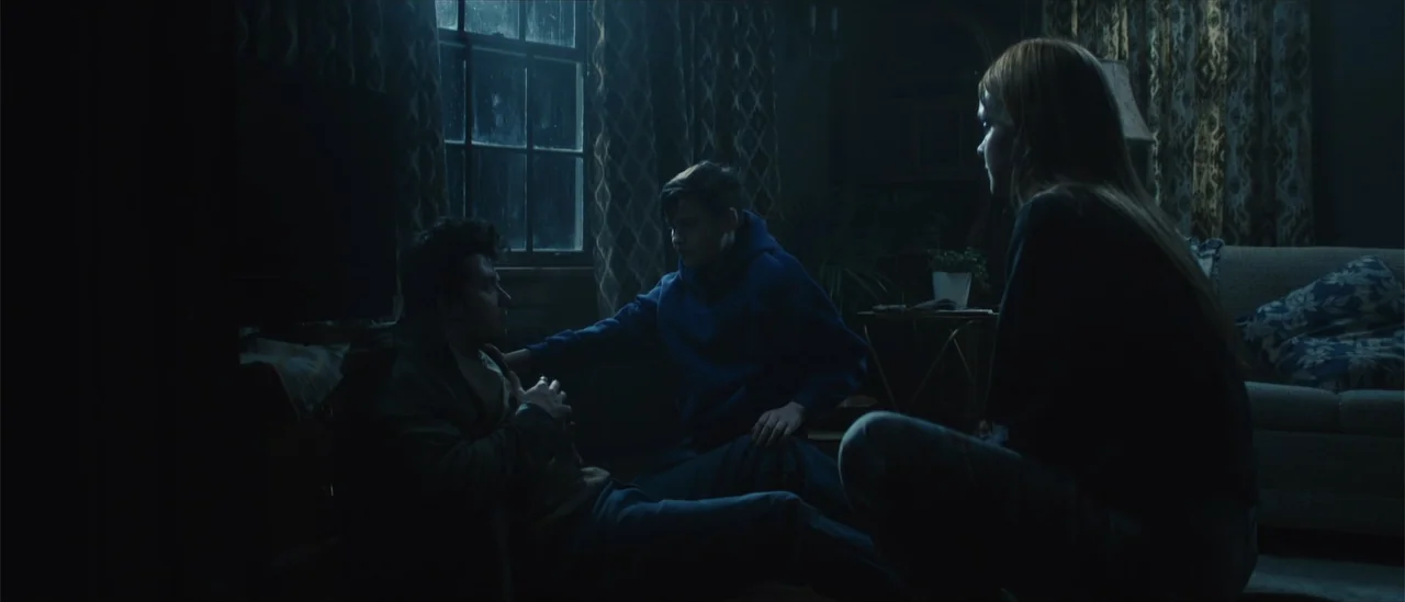 All Fun and Games (2023) movie still: medium — A dark, sparsely furnished space with heavy curtains and a rain-streaked window. A young boy in a…