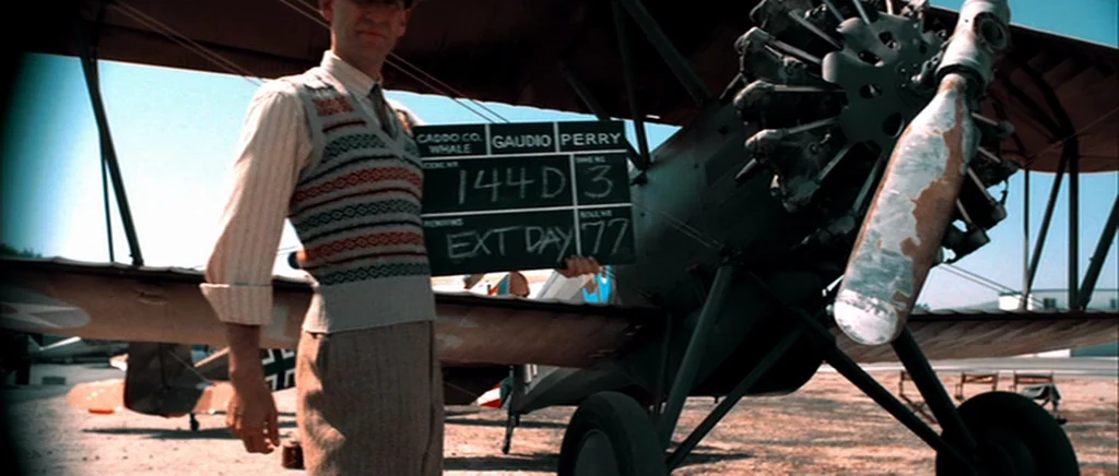 The Aviator (2004) movie still: medium — A sunny outdoor airfield features a vintage biplane parked on dry ground. A man holds a film clapperboard…