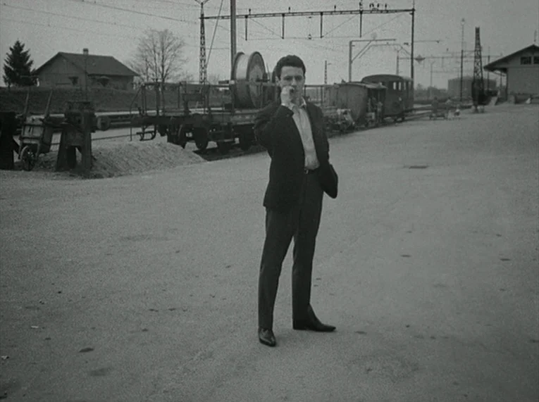 Le Petit Soldat (1963) movie still: fullbody — A desolate railway yard features industrial freight cars and overhead power lines. A young man…