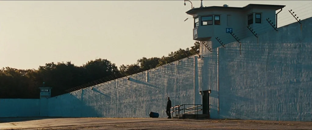 The Town (2010) movie still: wide — A high, whitewashed concrete wall with razor wire and a guard tower spans the frame. A single man stands alone…