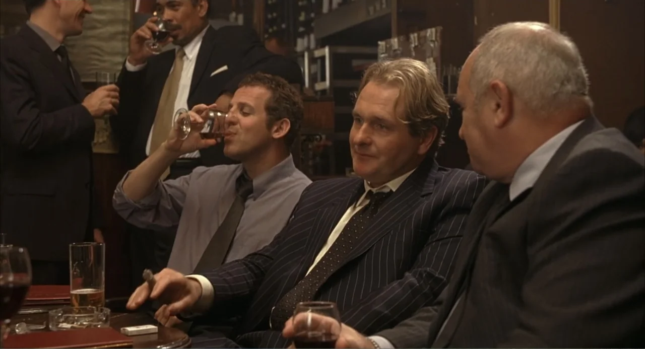 Scoop (2024) movie still: medium — A dimly lit restaurant interior with dark wood paneling and multiple tables. Three men in suits sit together at a…