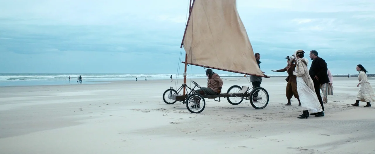 Slack Bay (2016) movie still: wide — A wide, flat sandy beach stretches toward a calm grey sea under overcast skies. A group of people in historical…