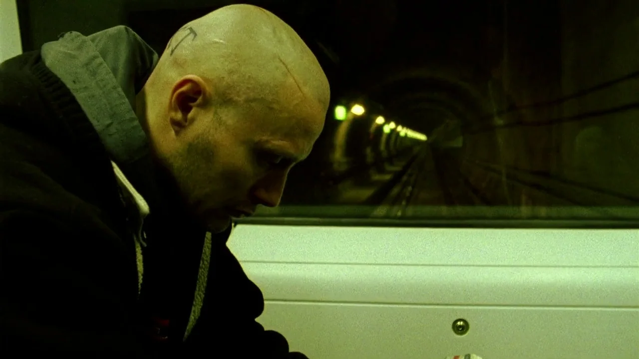 Pusher II (2004) movie still: medium — A bald man sits in a subway train traveling through a dark tunnel. The carriage interior is bathed in a…