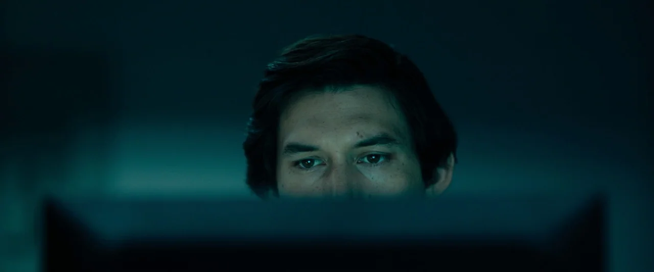 The Report (2019) movie still: closeup — A man looks at a screen in a dark room with minimal architectural detail. The light from the display casts…