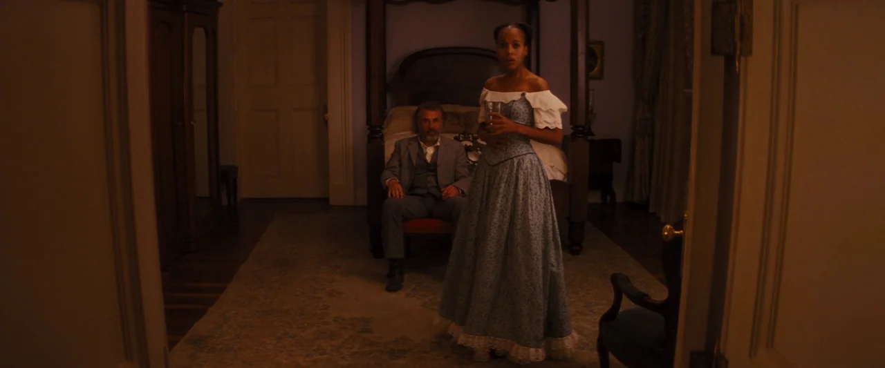 Django Unchained (2012) movie still: fullbody — A dimly lit 19th-century room featuring a large dark wood four-poster bed. A man in a suit sits in a…