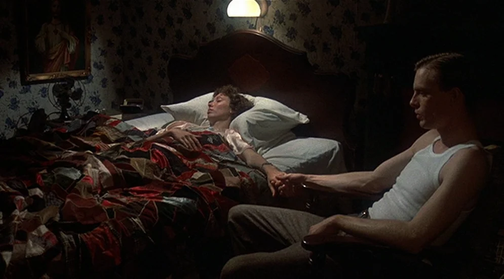 Thieves Like Us (1974) movie still: wide — A dark bedroom with floral wallpaper, a wooden bedframe, and a religious painting on wall. The man sits…