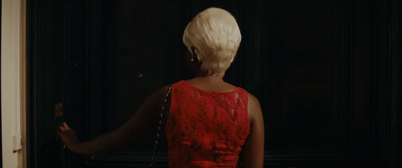 Girlhood (2014) movie still: medium — A dark, paneled door stands at the end of a corridor. A woman with blonde hair opens the door while wearing a…