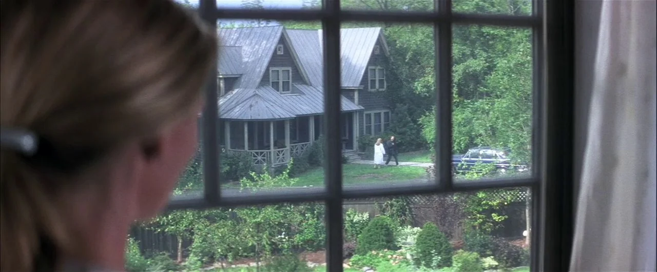 What Lies Beneath (2000) movie still: medium — A view through a window showing a two-story house with a porch. Two individuals stand on the walkway…