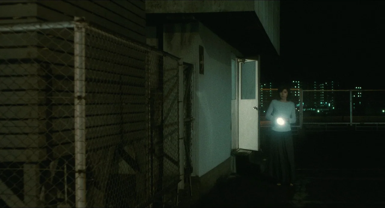 Dark Water (2002) movie still: wide — A dark night exterior features a chain-link fence beside a white building structure. A lone woman stands in…