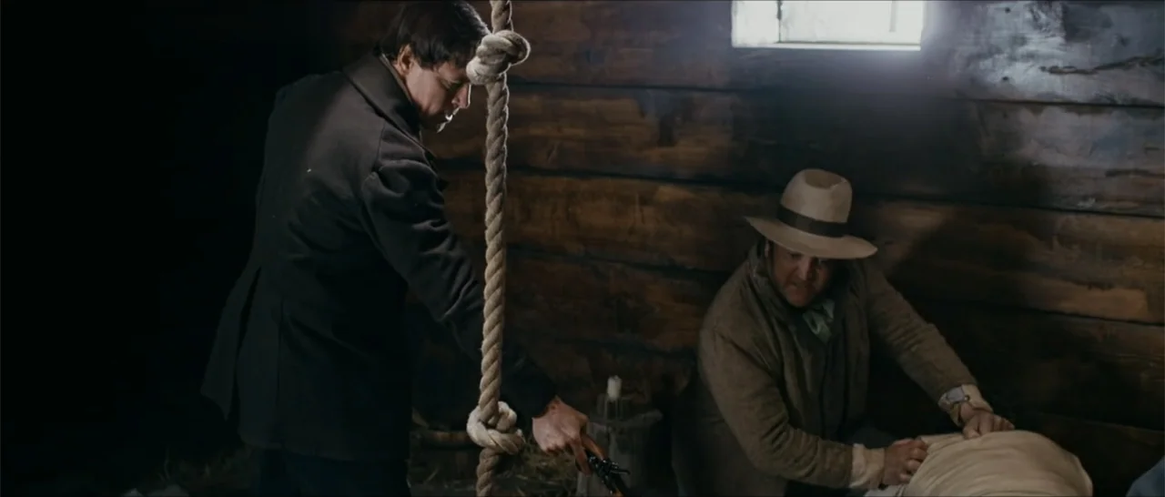 Out of Liberty (2019) movie still: medium — A dimly lit room with rustic dark wood walls and a rope hanging vertically. A man in a dark coat points…
