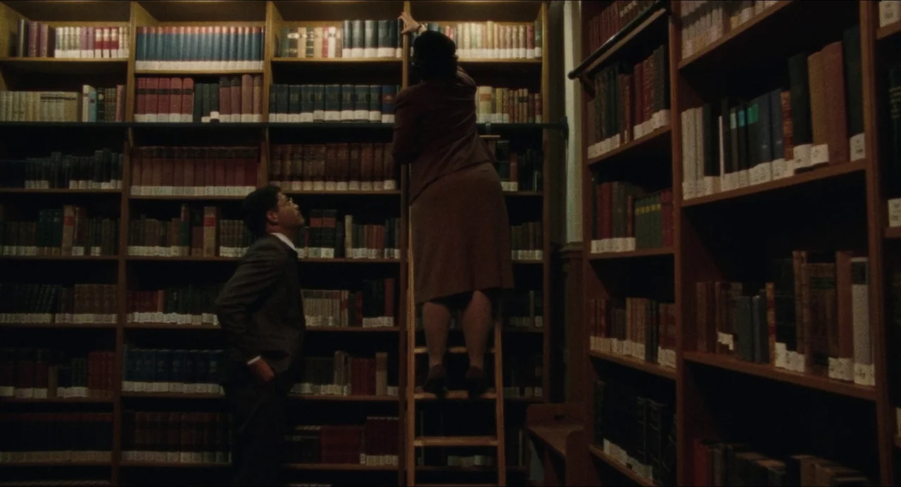 Origin (2023) movie still: medium — A dense room filled with tall, dark wooden shelving units packed with aged books. A woman on a wooden ladder…