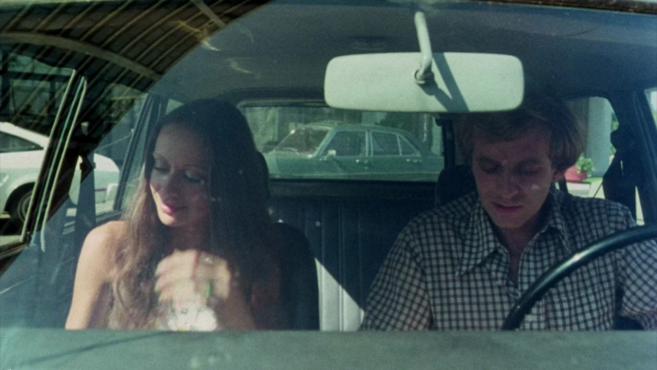 Shivers (1975) movie still: medium — The interior of a 1970s sedan with a dashboard and rearview mirror visible. A man operates the steering wheel…