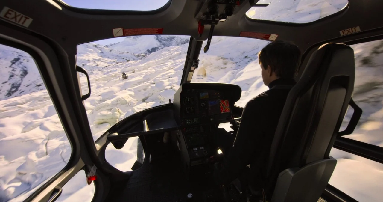 The interior of a helicopter cockpit displays modern flight instruments and large observation windows. The pilot controls the aircraft as it flies over a vast expanse of snow.