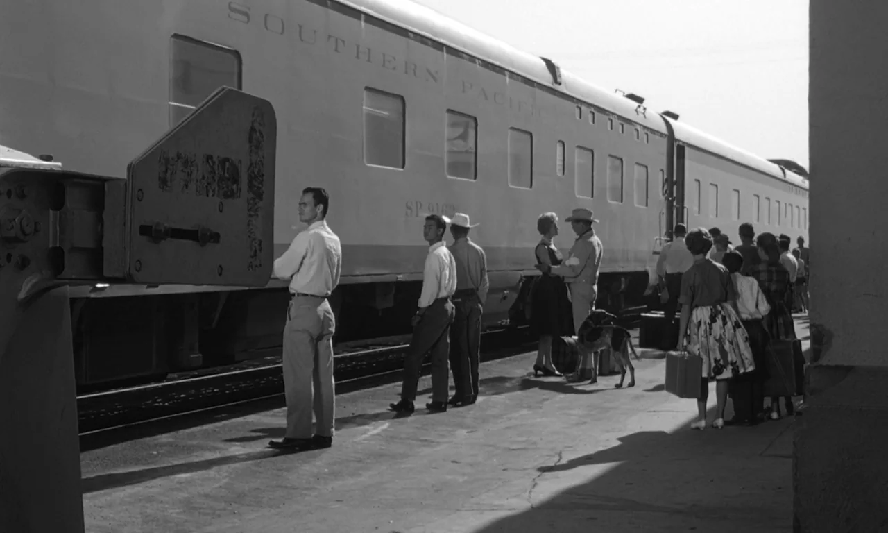 The Misfits (1961) movie still: wide — A sunny concrete train platform features a large Southern Pacific passenger train car. Several people stand…