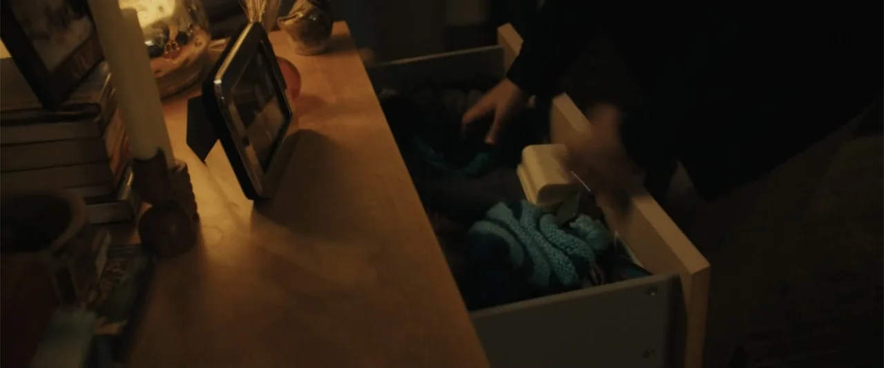 Afraid (2024) movie still: medium — A wooden chest of drawers sits in a dimly lit room with personal items atop. An individual reaches into the…