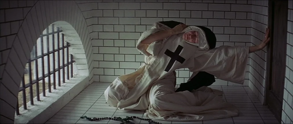 The Devils (1971) movie still: fullbody — A stone cell with white square tiles on the floor and walls. A nun kneels on the ground near a barred…