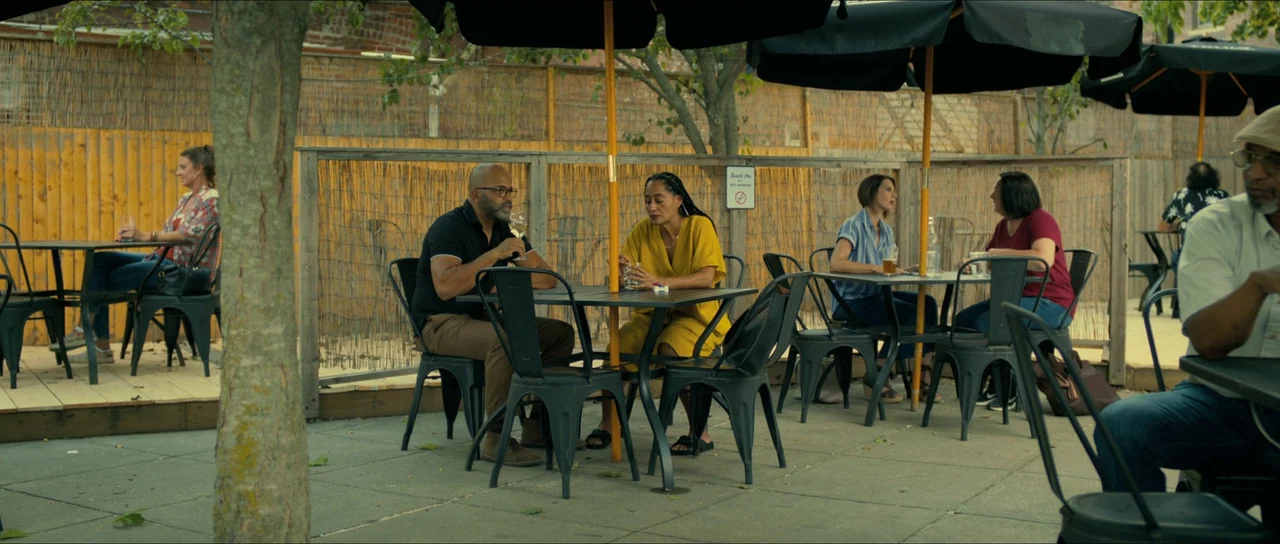 American Fiction (2023) movie still: wide — A patio cafe area features metal tables and chairs under black umbrellas. Several people sit at separate…