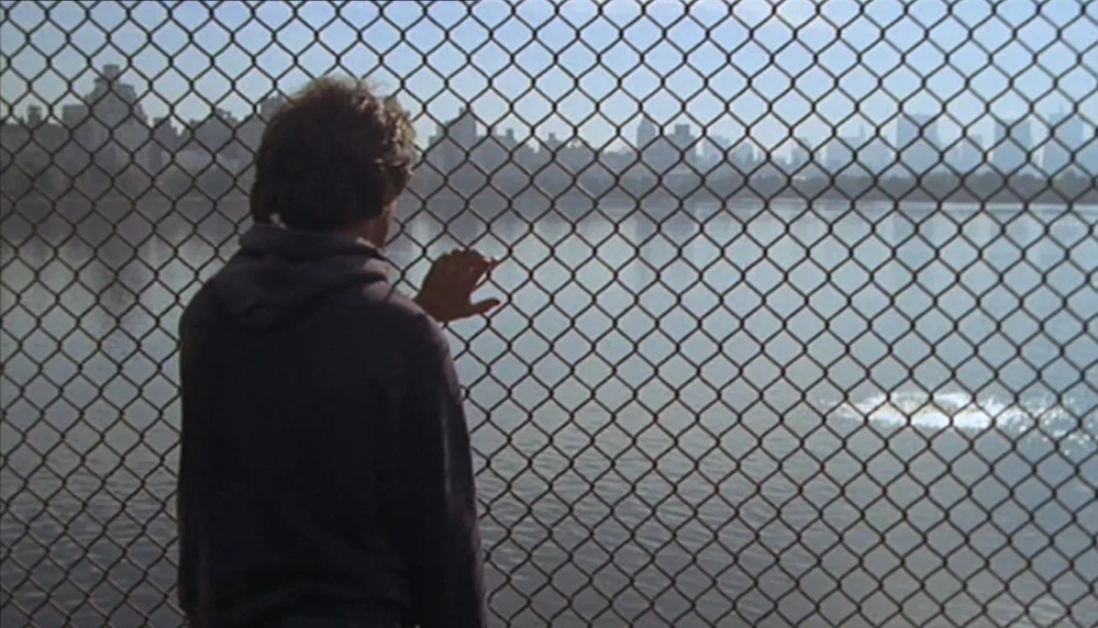 Marathon Man (1976) movie still: medium — A man stands behind a chain-link fence overlooking a large urban reservoir. The distant city skyline is…
