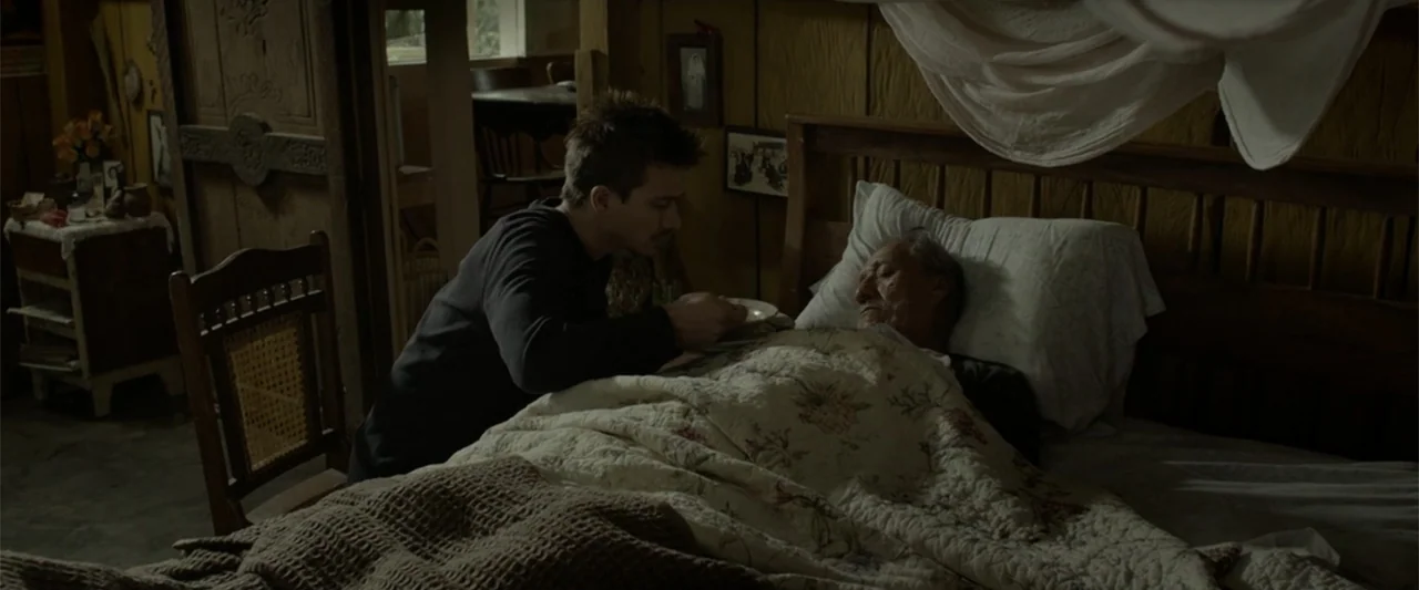 I Dream in Another Language (2017) movie still: medium — A wooden interior room contains a bed with a floral quilt and pillows. A young man sits at…