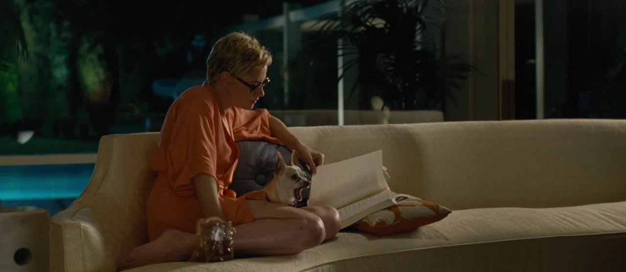 Seberg (2019) movie still: medium — A woman in a robe sits on a light-colored sofa at night. She holds a book open while a small dog rests on her lap.