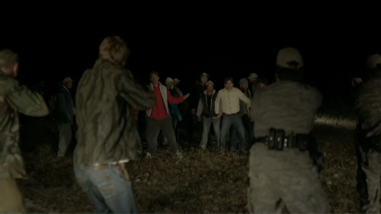 Loco Love (2003) movie still: wide — A dark, uneven field at night is lit by bright, low-angle light. A group of men stands in the open while others…