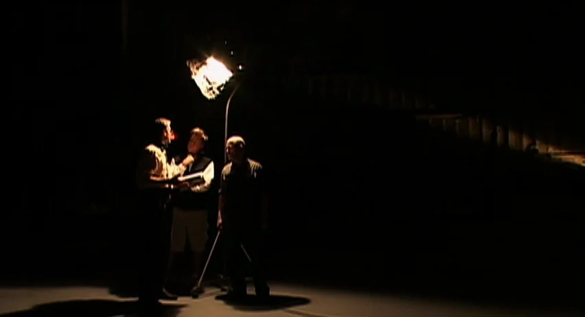 Inland Empire (2006) movie still: wide — A dark, empty space illuminated by a single harsh overhead spotlight. Three men are standing in the center…