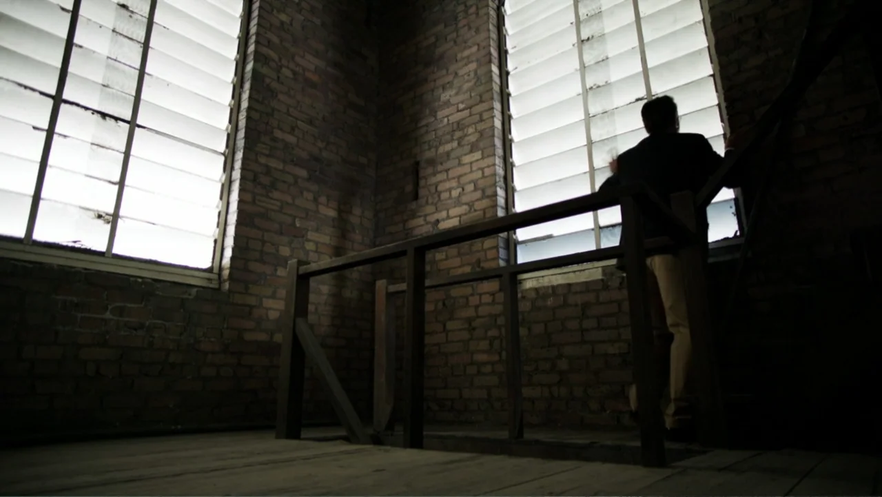 At Middleton (2013) movie still: medium — A dark brick stairwell landing features large windows with horizontal blinds and wooden railings. A man in…