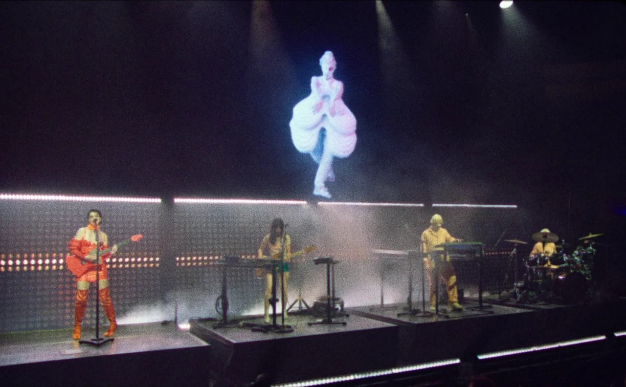 The Nowhere Inn (2020) movie still: wide — A dark concert stage features four musicians positioned behind a large illuminated screen. A holographic…