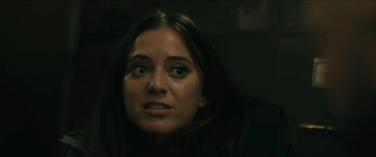 Guns Up (2025) movie still: closeup — A dimly lit interior space with a blurred, dark background. A young woman focuses her gaze on an unseen person…