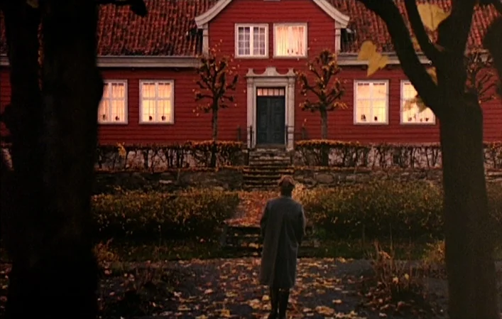 Autumn Sonata (1978) movie still: wide — A red wooden manor stands behind a stone wall and formal landscape of fallen leaves. A man in a grey coat…