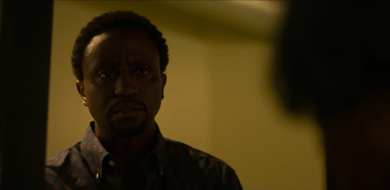 Kimi (2022) movie still: closeup — An dimly lit interior space with shadows obscuring the surroundings. A man looks toward someone standing just out…