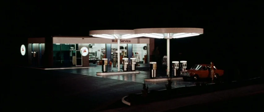 Messiah of Evil (1973) movie still: establishing — A 1970s Mobil gas station with round canopy lights sits in a dark landscape. The pumps and…