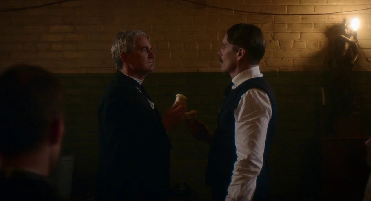 Tesla (2020) movie still: medium — A dimly lit room features a painted brick wall with an exposed practical light fixture. Two men in formal 1940s…
