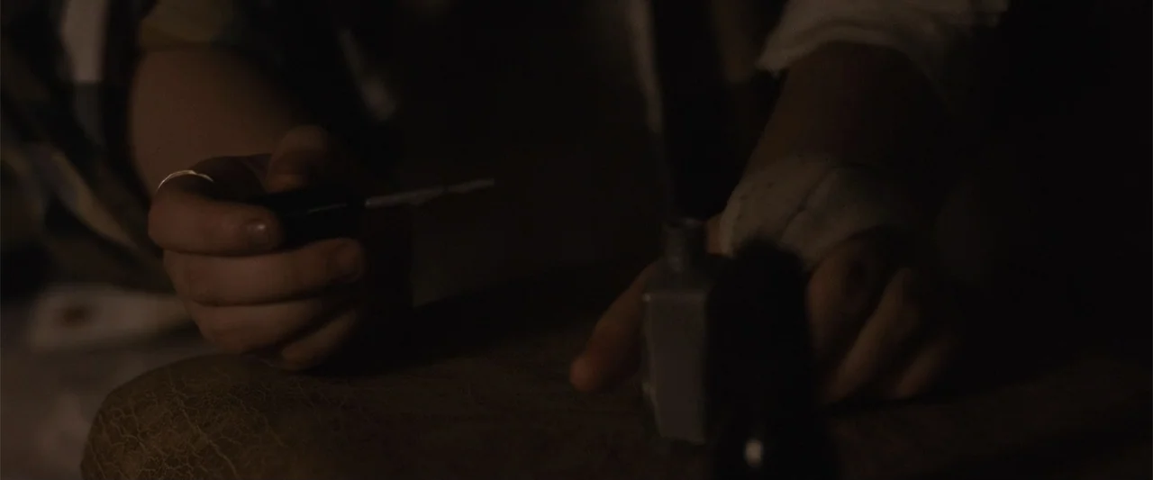 Maggie (2015) movie still: closeup — A dimly lit interior space features hands performing a precise task over a surface. The hands hold an…