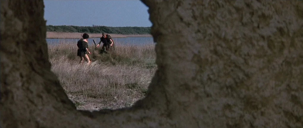 Fellini – Satyricon (1969) movie still: wide — Two figures with swords stand in a marshy landscape near a blue lake. A textured, irregular opening…
