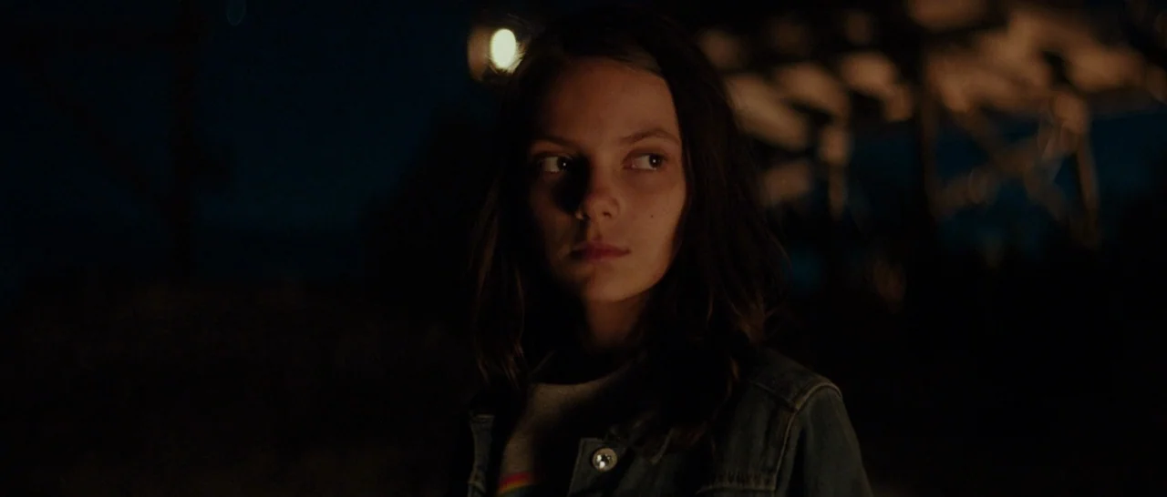 Logan (2017) movie still: closeup — A girl stands outdoors in a dark, rural environment at night. She looks toward the side of the frame while lit…