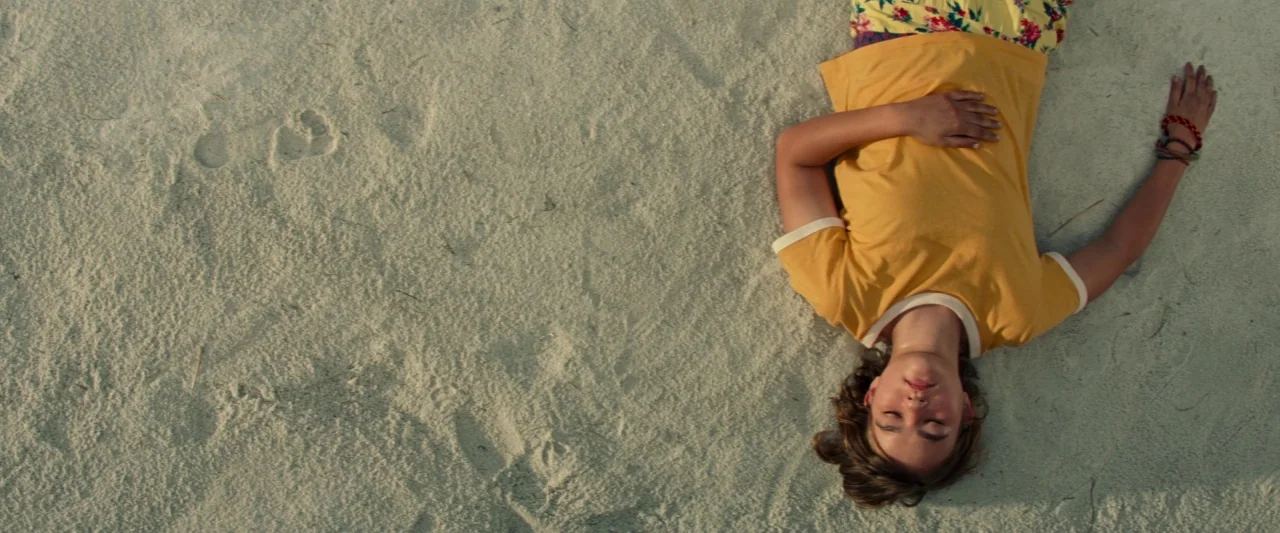 A Quiet Place (2018) movie still: wide — A person lies on pale, textured sand under direct daylight. The individual remains motionless on the sand…