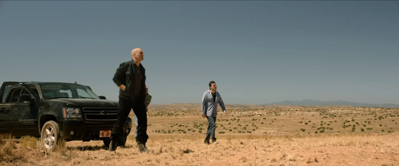 The Marksman (2021) movie still: fullbody — A vast, arid desert landscape features dry brush and distant mountains under a clear sky. Two men stand…