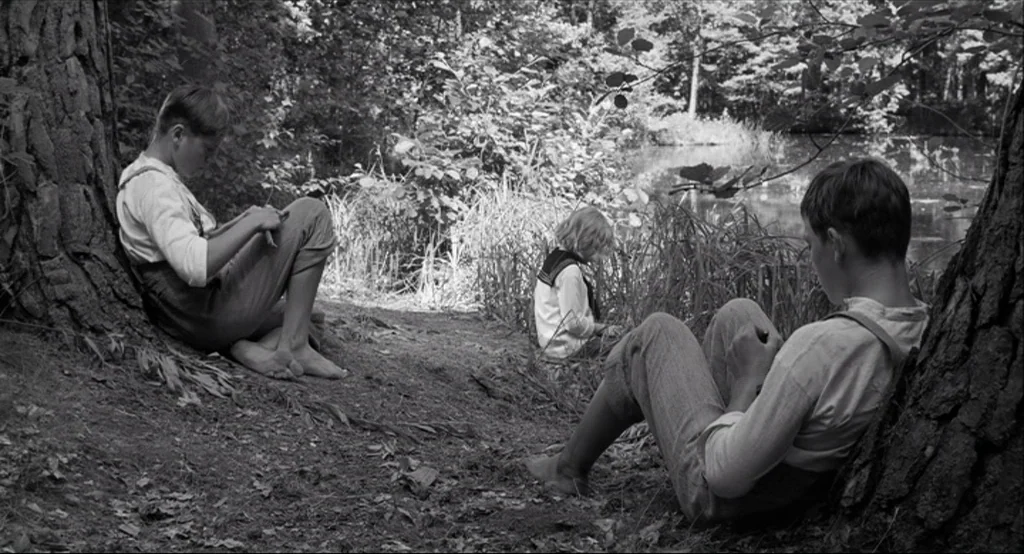The White Ribbon (2009) movie still: wide — A rural lakeshore area with dense trees and long grass banks. Three children sit quietly on the dirt…