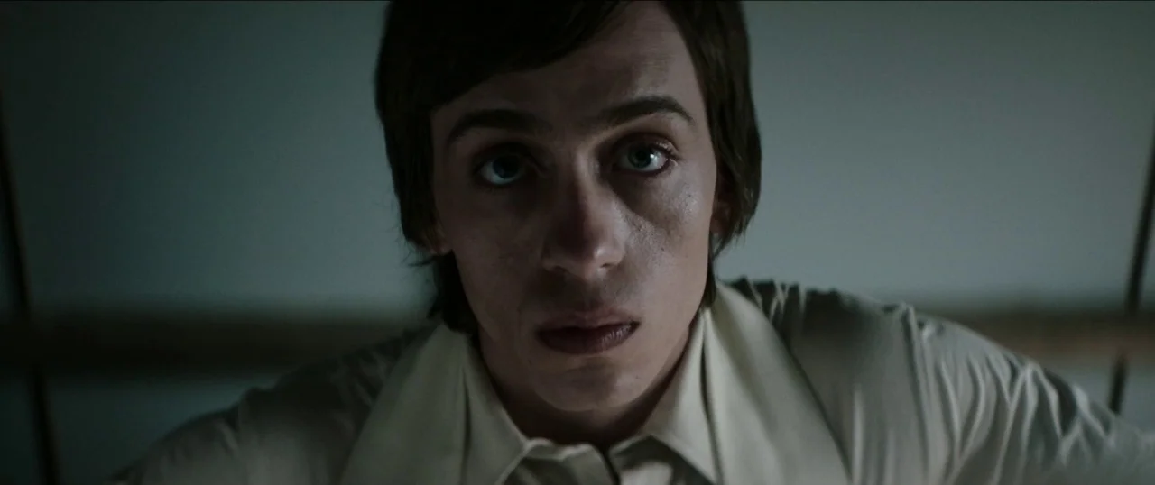 Good Favour (2017) movie still: closeup — A dark indoor space with a plain neutral background. A young man stares directly at the camera.
