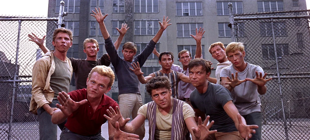 West Side Story (1961) movie still: medium — A group of men stand before a chain-link fence and a tall brick building. They hold their hands out…